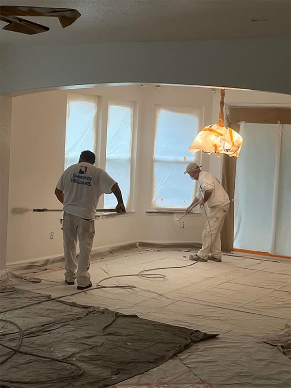 Two Bayshore Painters employees painting interior walls with protective tarps covering floors and windows